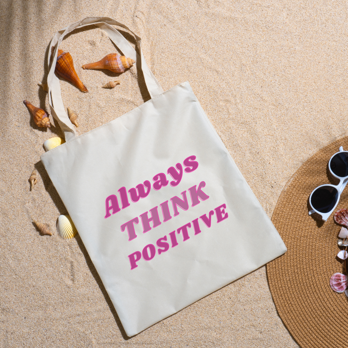 Always Think Positive tote on beach sand ringed with seashells beside a straw hat and sunglasses