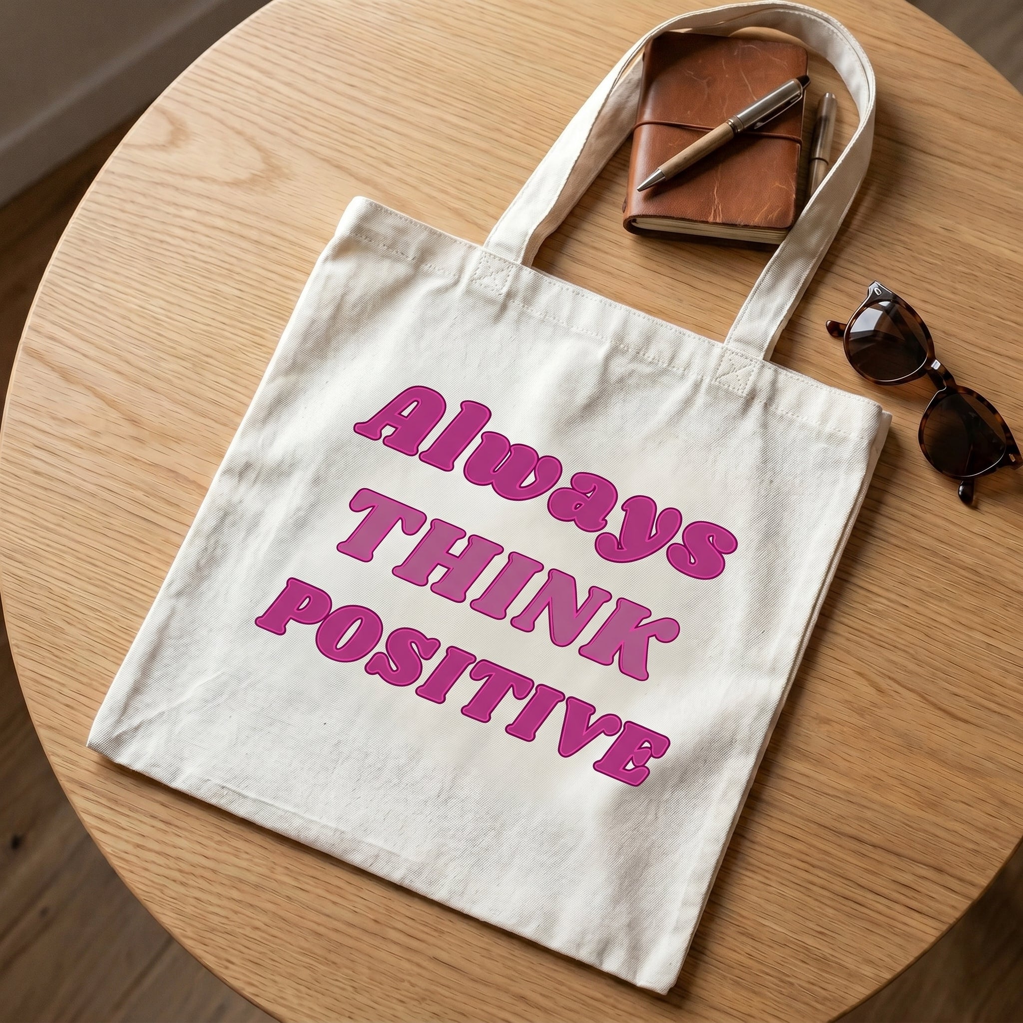 Always Think Positive tote on a round oak table with a tan leather notebook, two pens and sunglasses