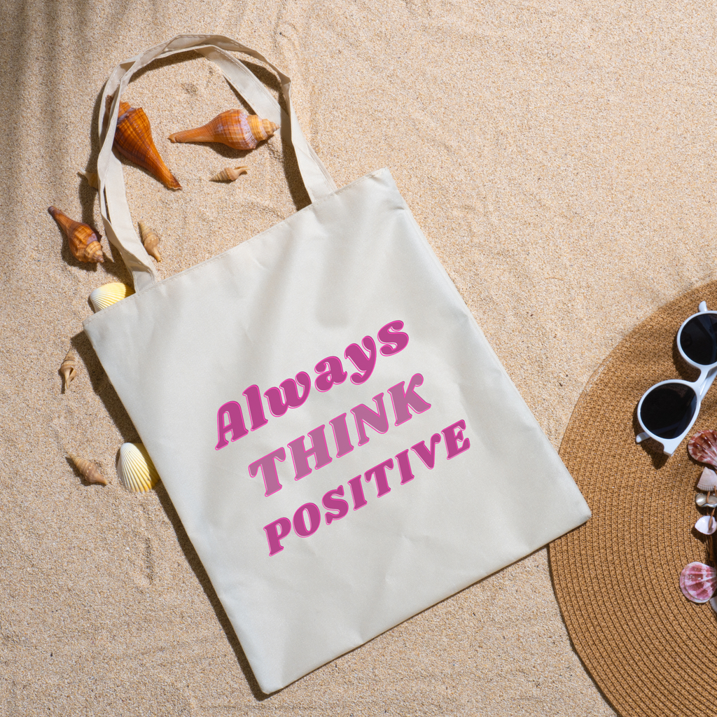 Always Think Positive tote on beach sand ringed with seashells beside a straw hat and sunglasses