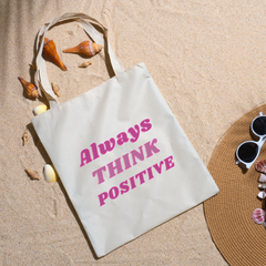 Always Think Positive tote on beach sand ringed with seashells beside a straw hat and sunglasses