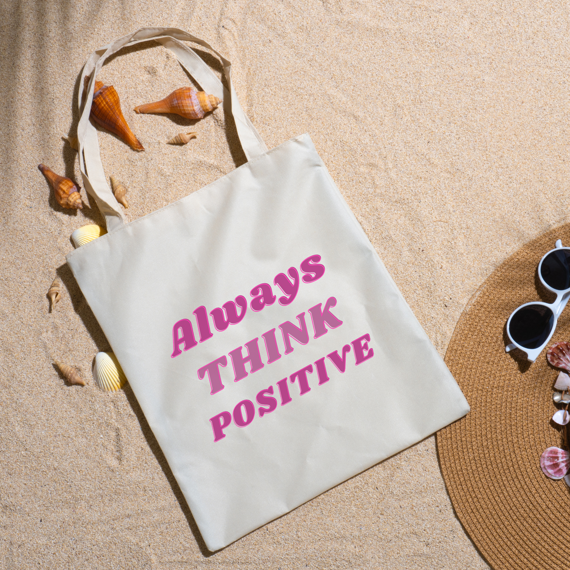 Always Think Positive tote on beach sand ringed with seashells beside a straw hat and sunglasses