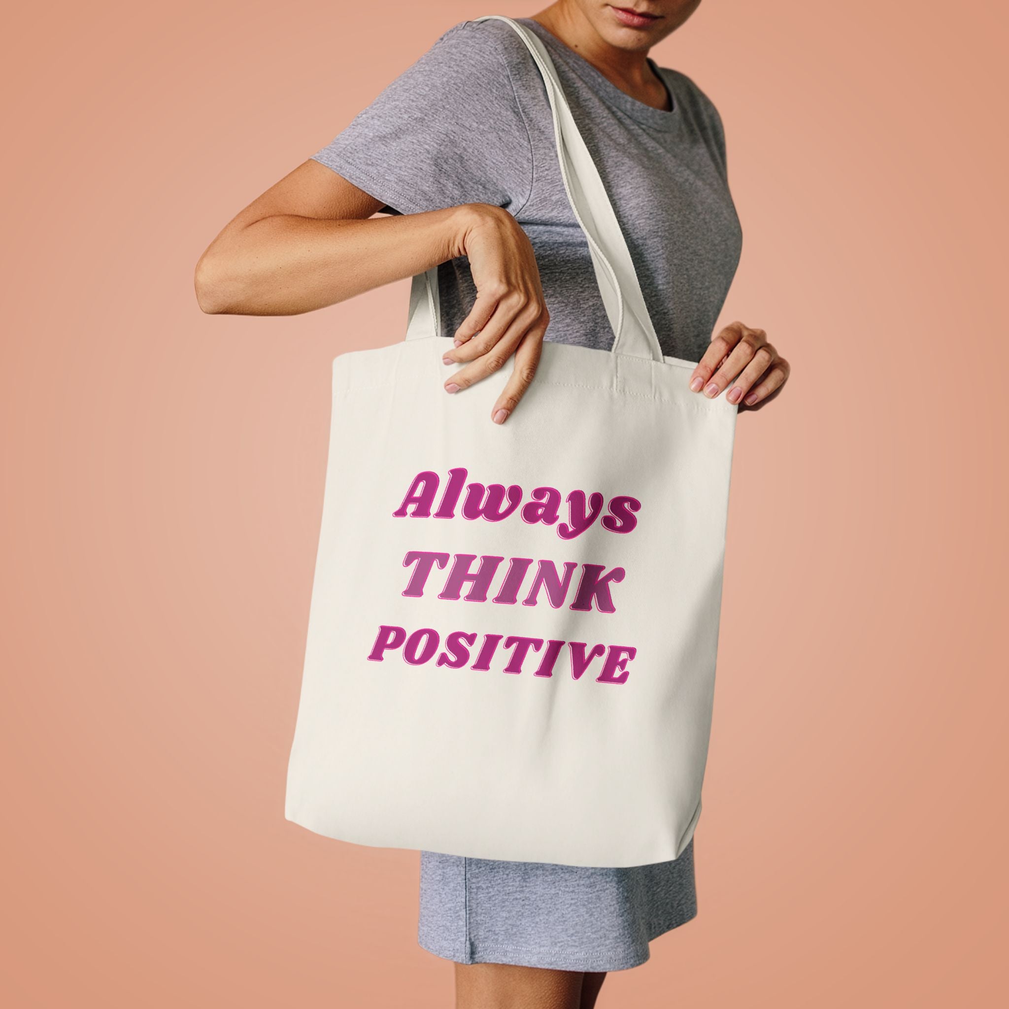 Woman in a grey tee lifting the Always Think Positive tote by its handles against peach