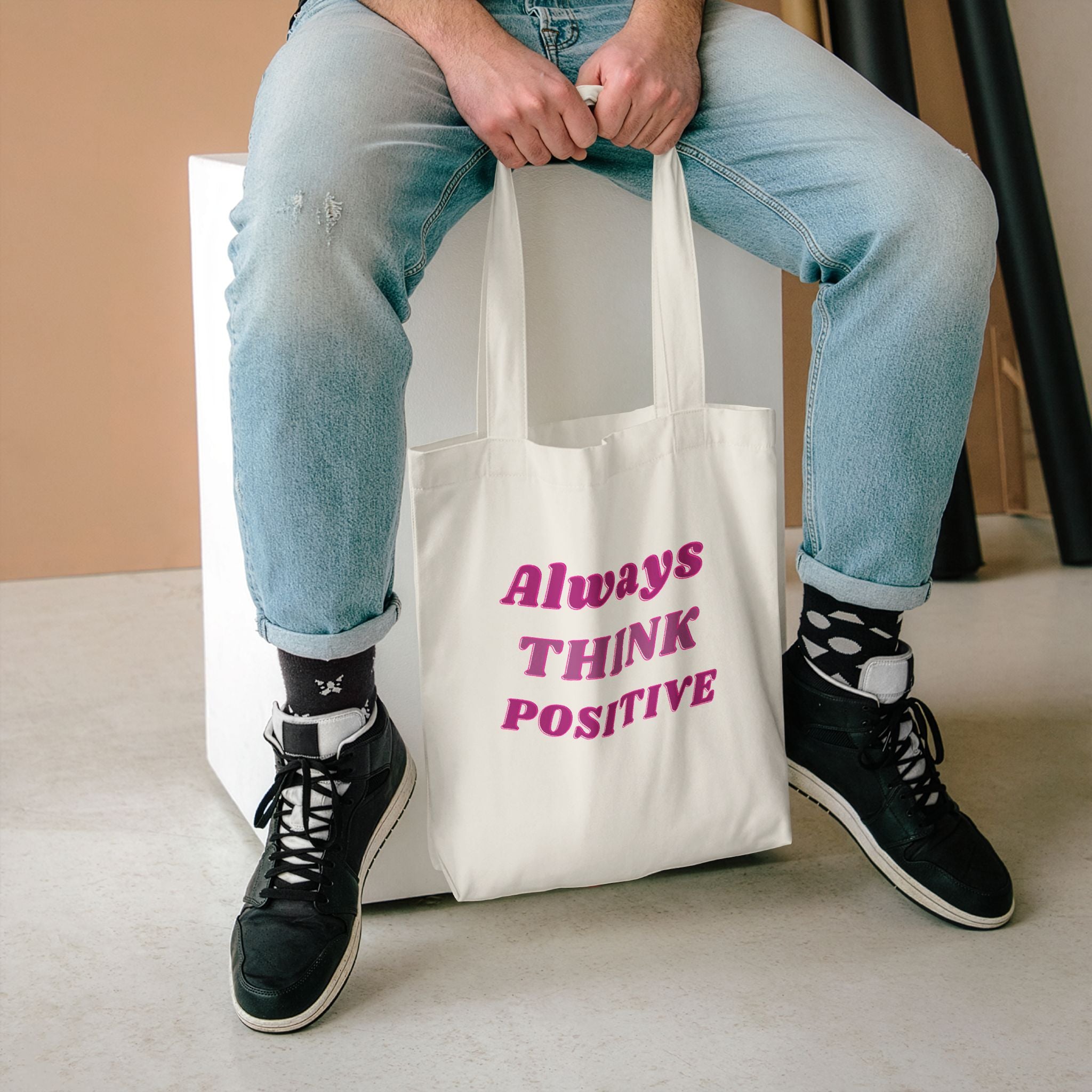 Always Think Positive tote held between the knees of someone seated in light jeans and high-tops