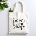 Plovera Co Born To Shop tote laid flat on pale grey with white blossom and green leaves
