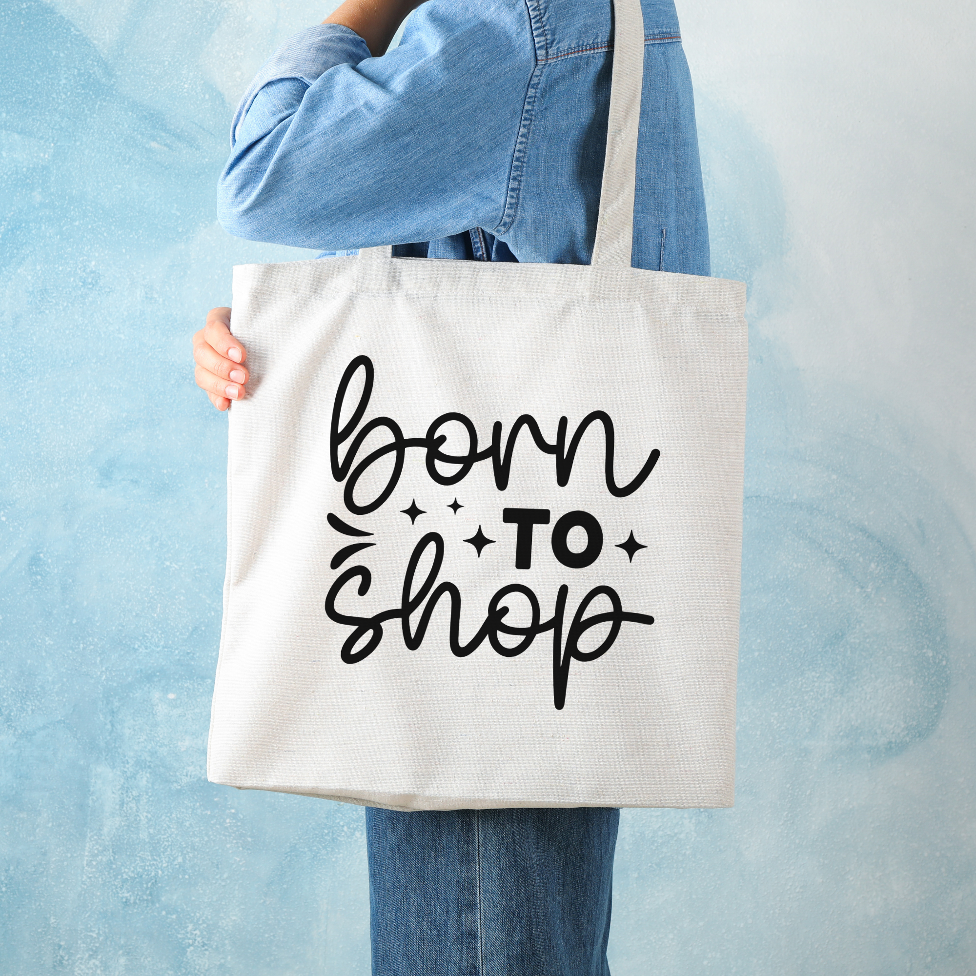 Person in a denim shirt carrying the Born To Shop tote against a mottled pale blue wall