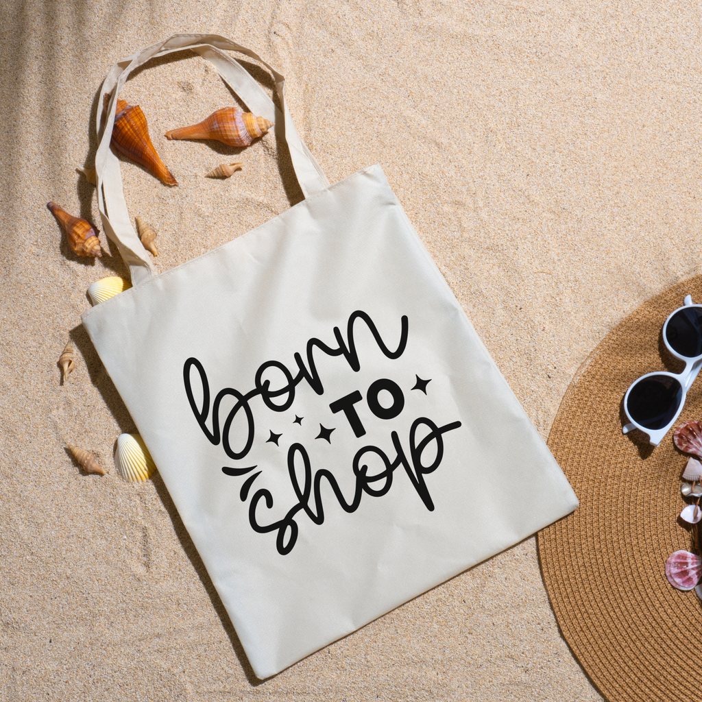Born To Shop tote on beach sand with seashells, a straw hat and white sunglasses nearby
