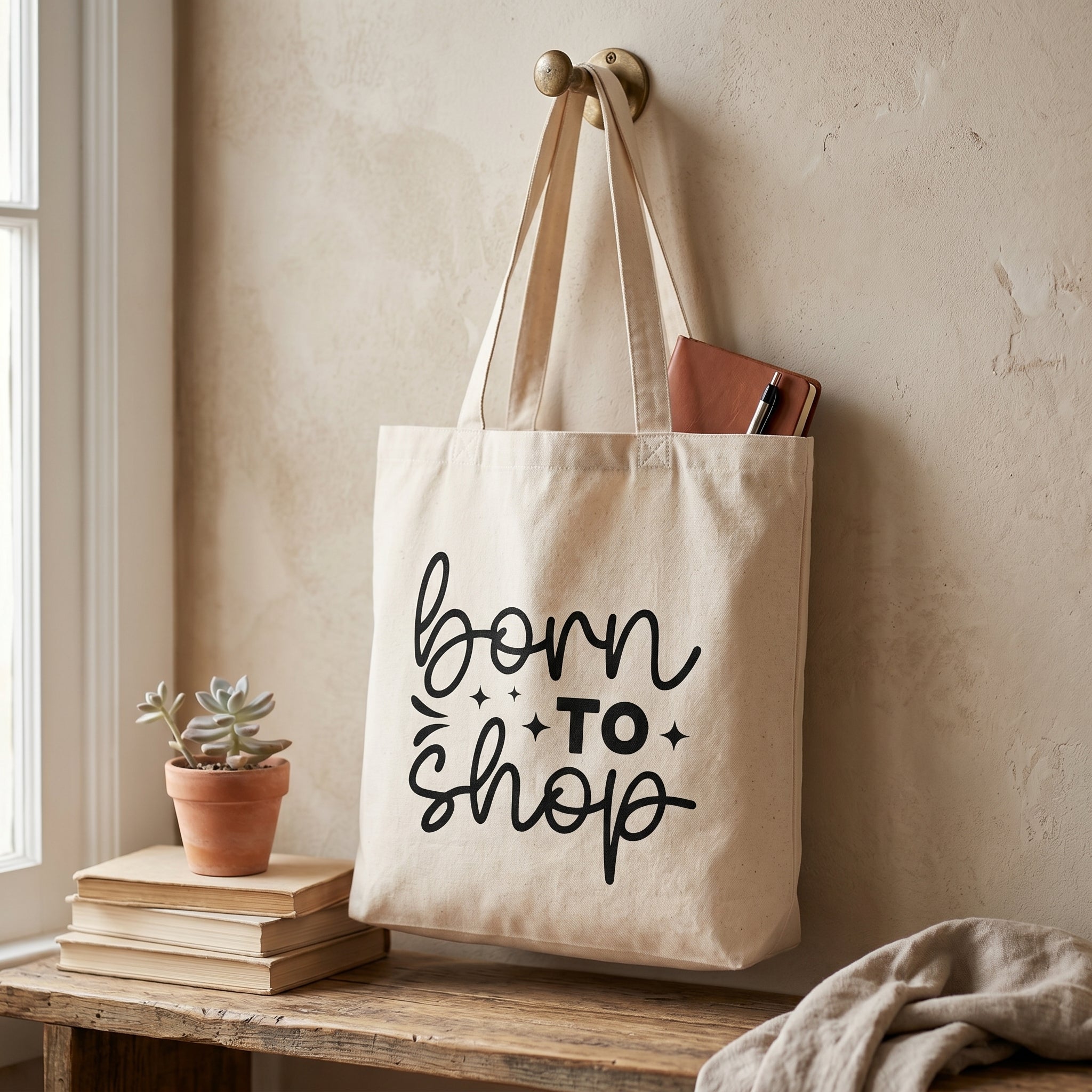 Born To Shop tote hanging from a brass hook with a notebook inside, succulent on books below