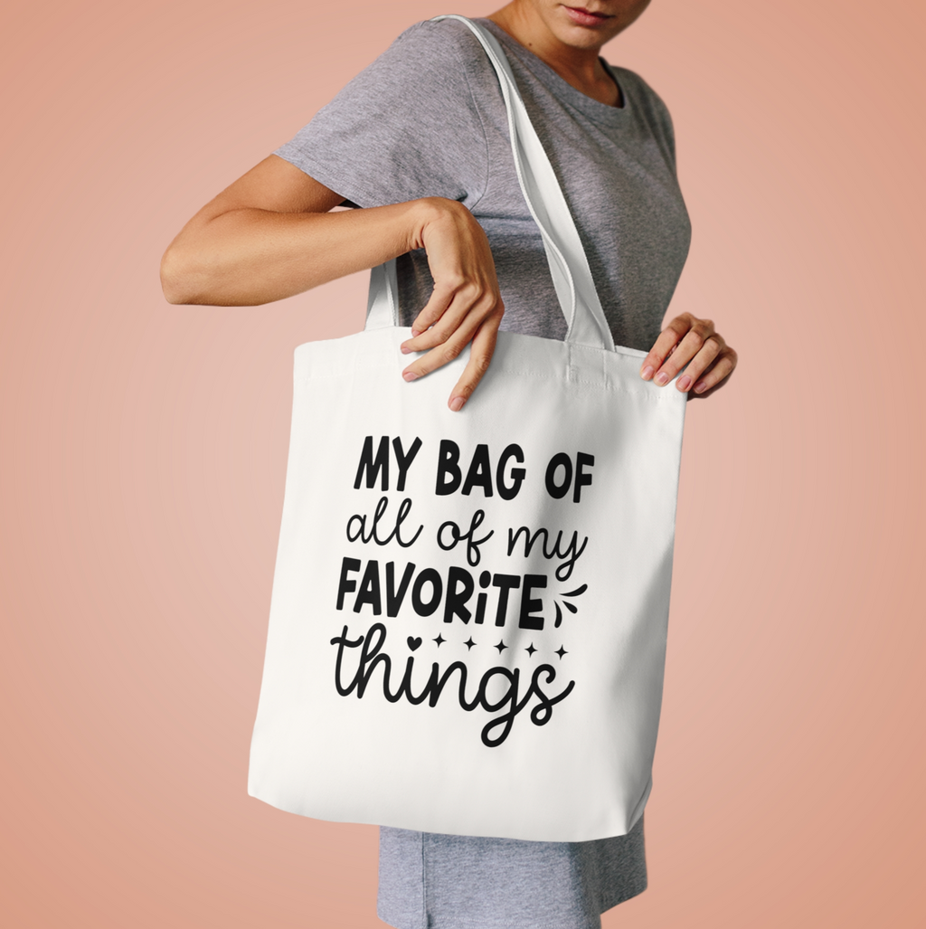 Woman in a grey tee lifting the Favorite Things tote by its handles against peach