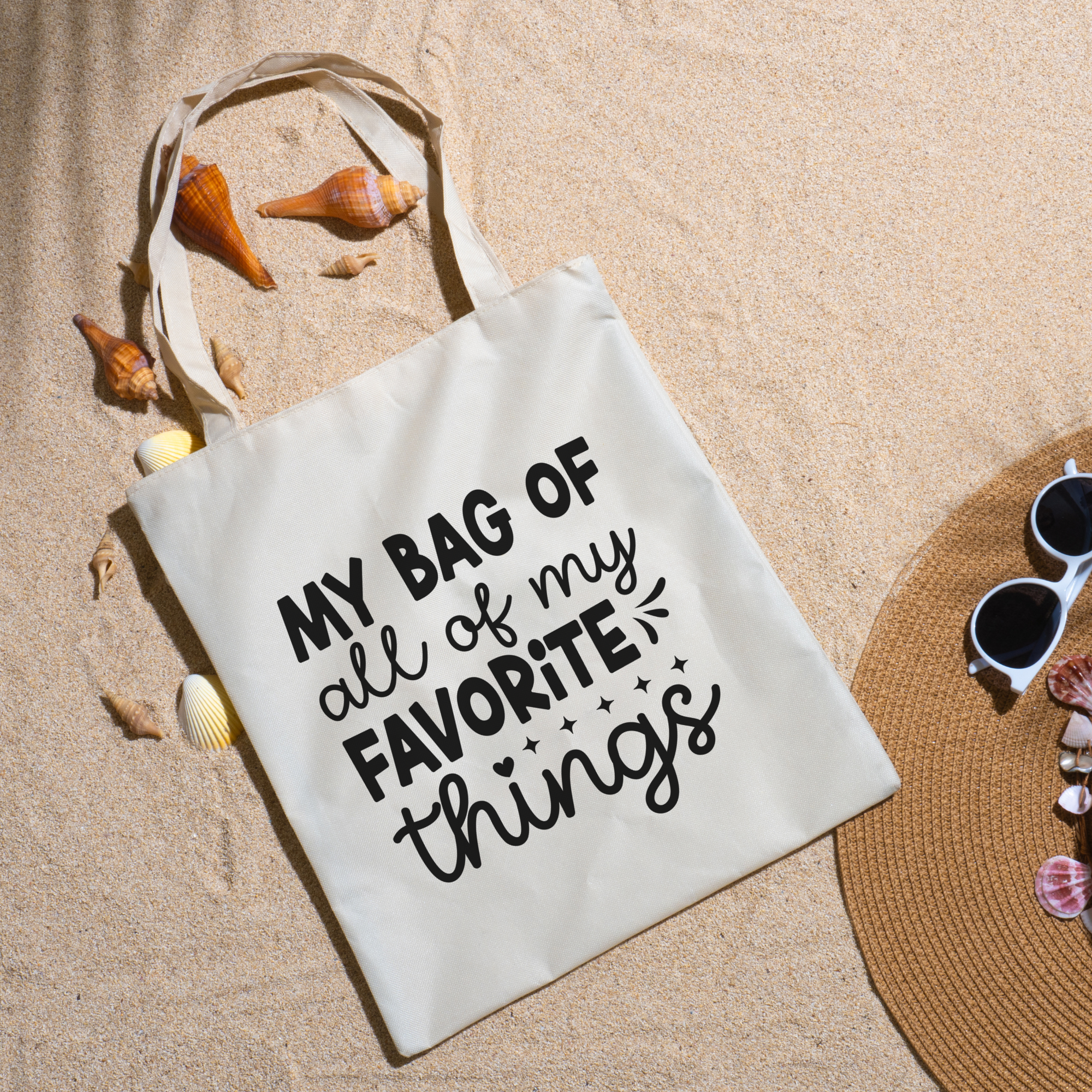 Favorite Things tote on beach sand with seashells, a straw hat and white sunglasses nearby