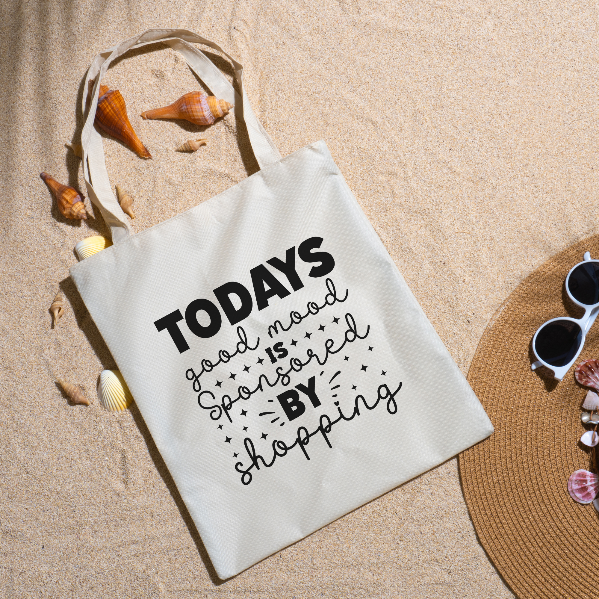 Good Mood tote on beach sand with seashells, a straw hat and white sunglasses nearby