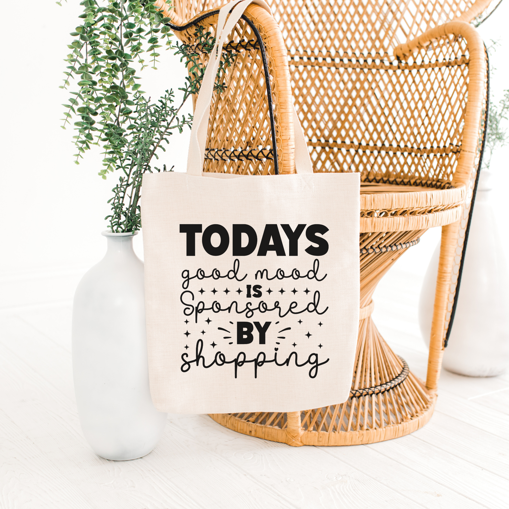 Good Mood tote propped against a rattan peacock chair beside a white vase of eucalyptus