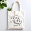 Plovera Co Enjoy the Little Things tote laid flat on pale grey with white blossom and green leaves