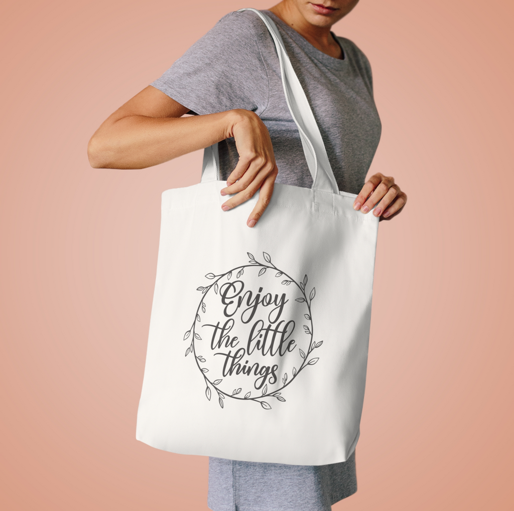 Woman in a grey tee lifting the Enjoy the Little Things tote by its handles against peach