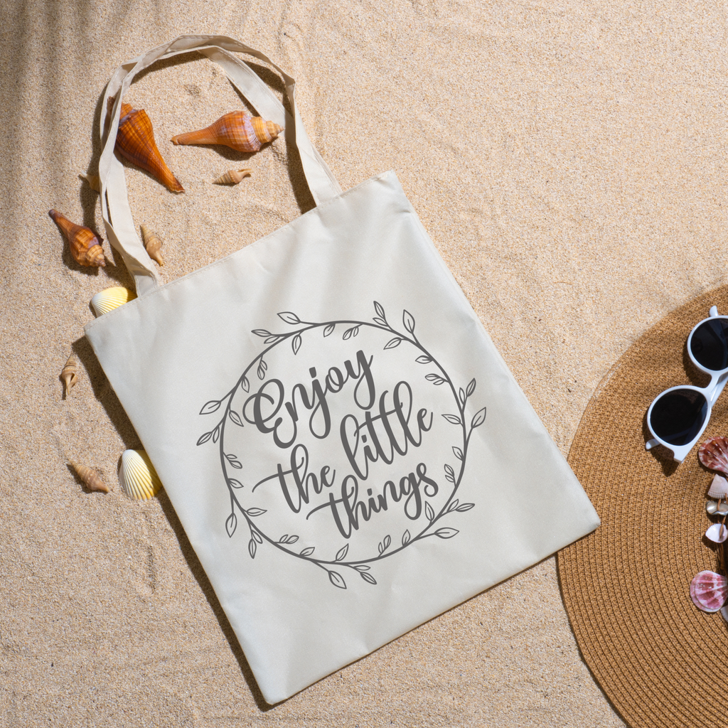 Enjoy the Little Things tote on beach sand with seashells, a straw hat and white sunglasses nearby