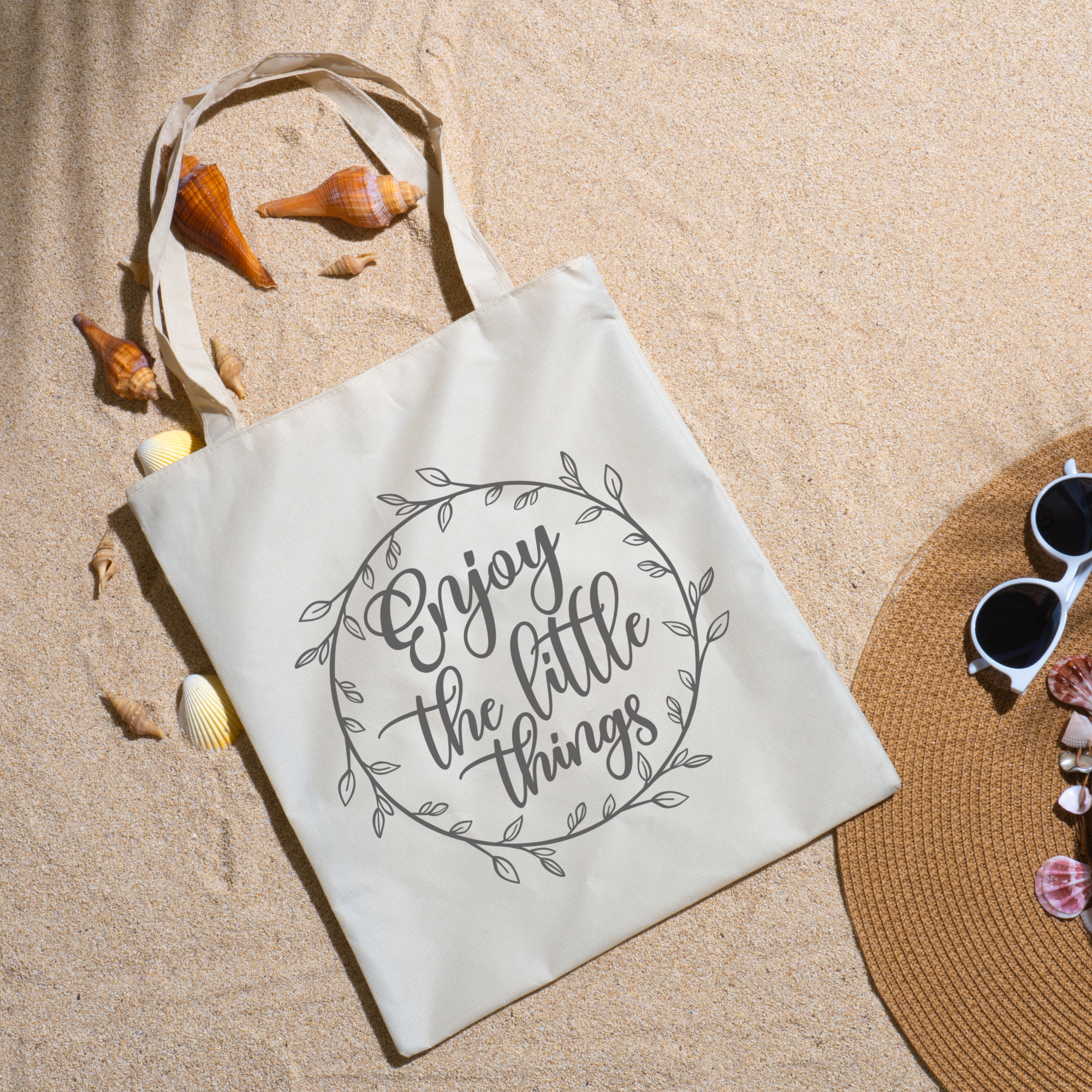 Enjoy the Little Things tote on beach sand with seashells, a straw hat and white sunglasses nearby