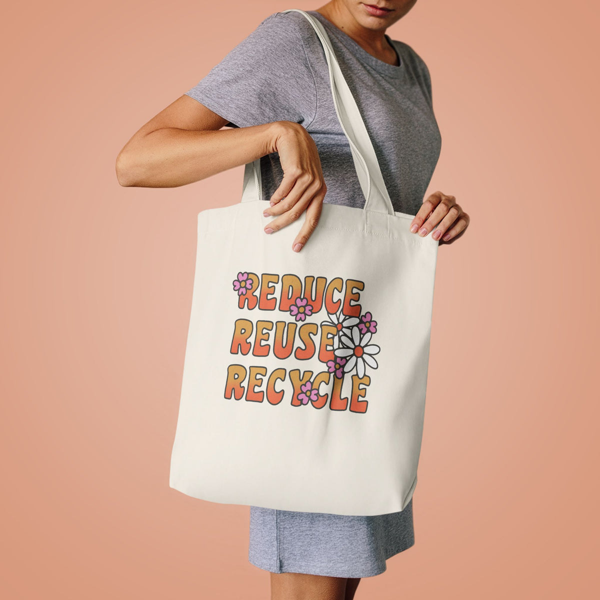 Woman in a grey tee lifting the Reduce Reuse Recycle tote by its handles against peach