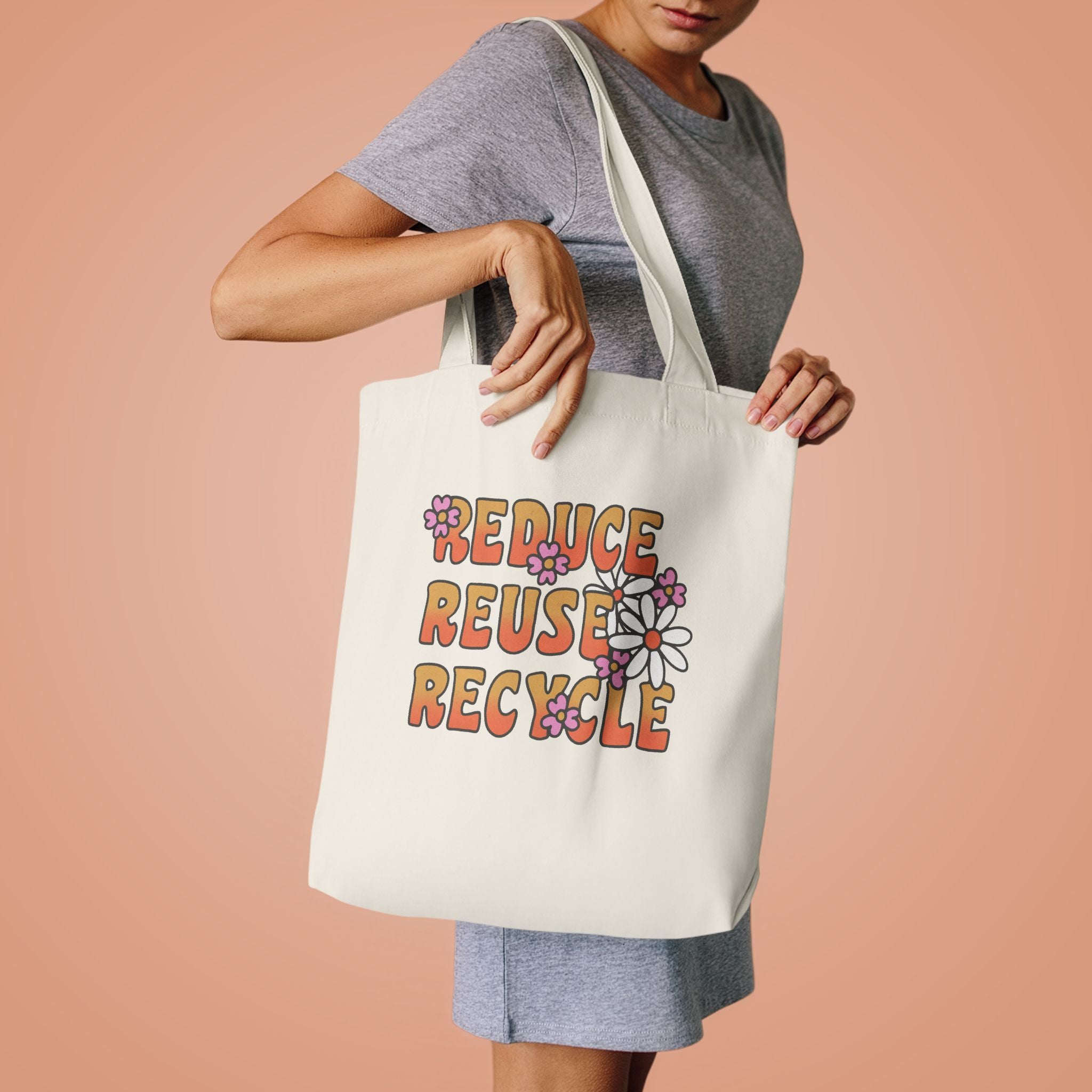 Woman in a grey tee lifting the Reduce Reuse Recycle tote by its handles against peach