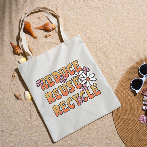 Reduce Reuse Recycle tote on beach sand with seashells, a straw hat and white sunglasses nearby