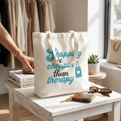 Shopping Is Cheaper Than Therapy tote on a white boutique side table beside keys and sunglasses