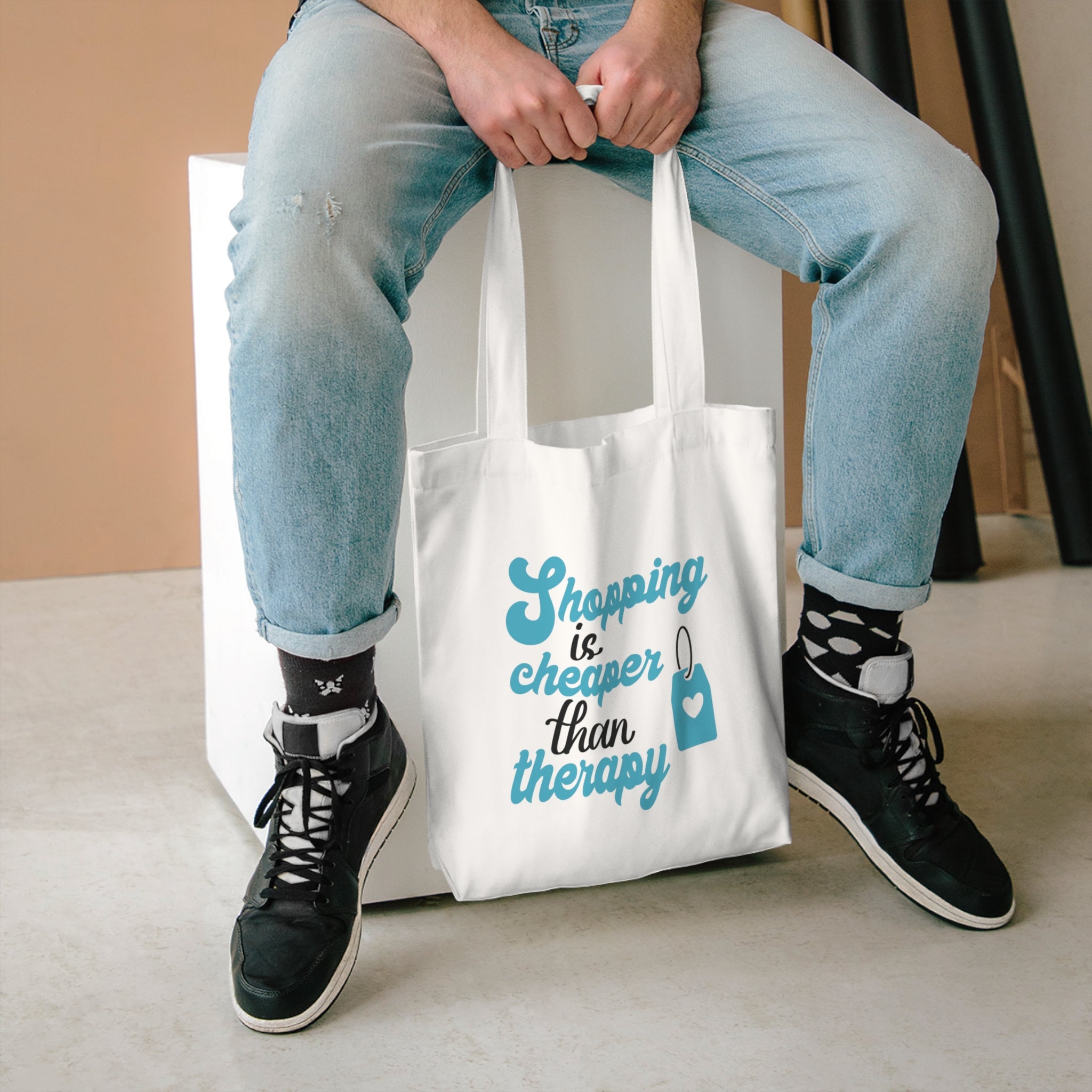 Shopping Is Cheaper Than Therapy tote held between the knees of someone seated in light jeans
