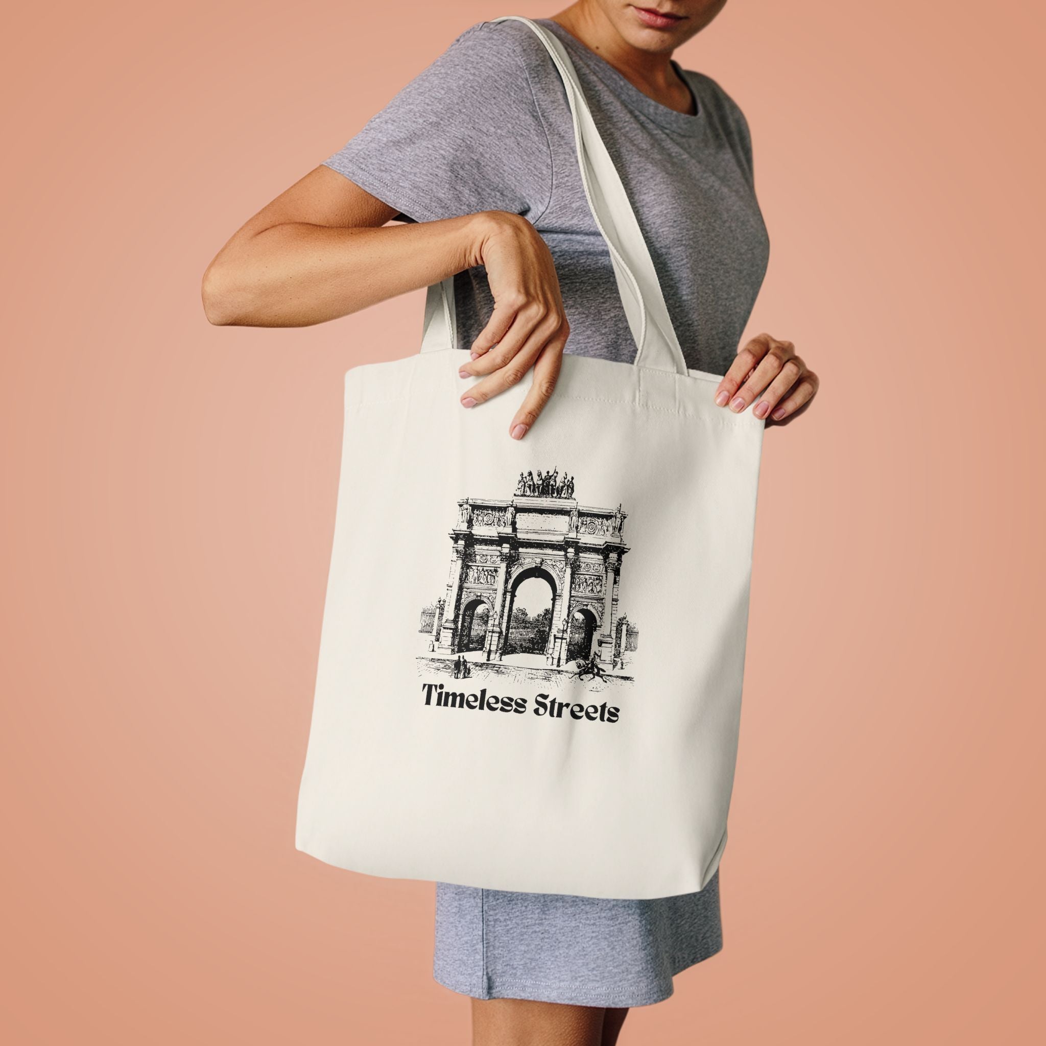 Woman in a grey tee lifting the Timeless Streets tote by its handles against peach