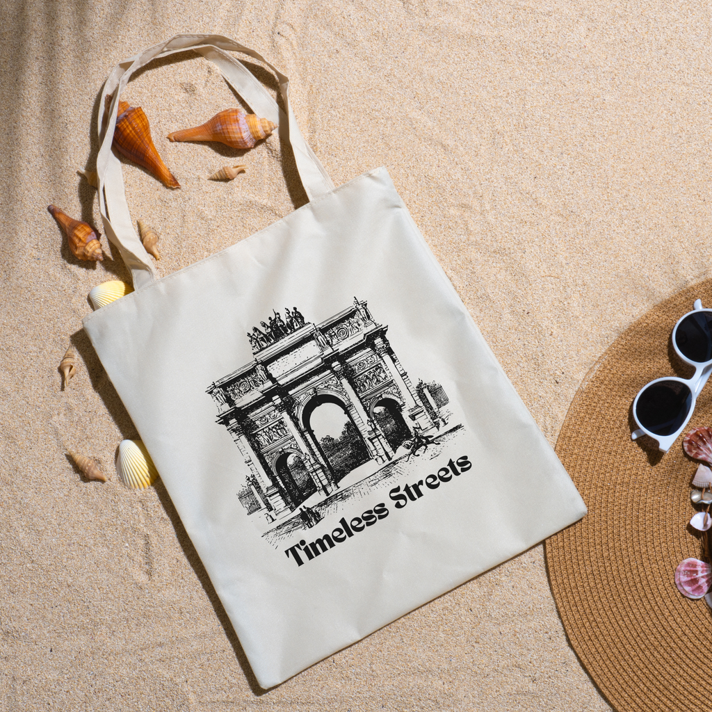 Timeless Streets tote on beach sand with seashells, a straw hat and white sunglasses nearby