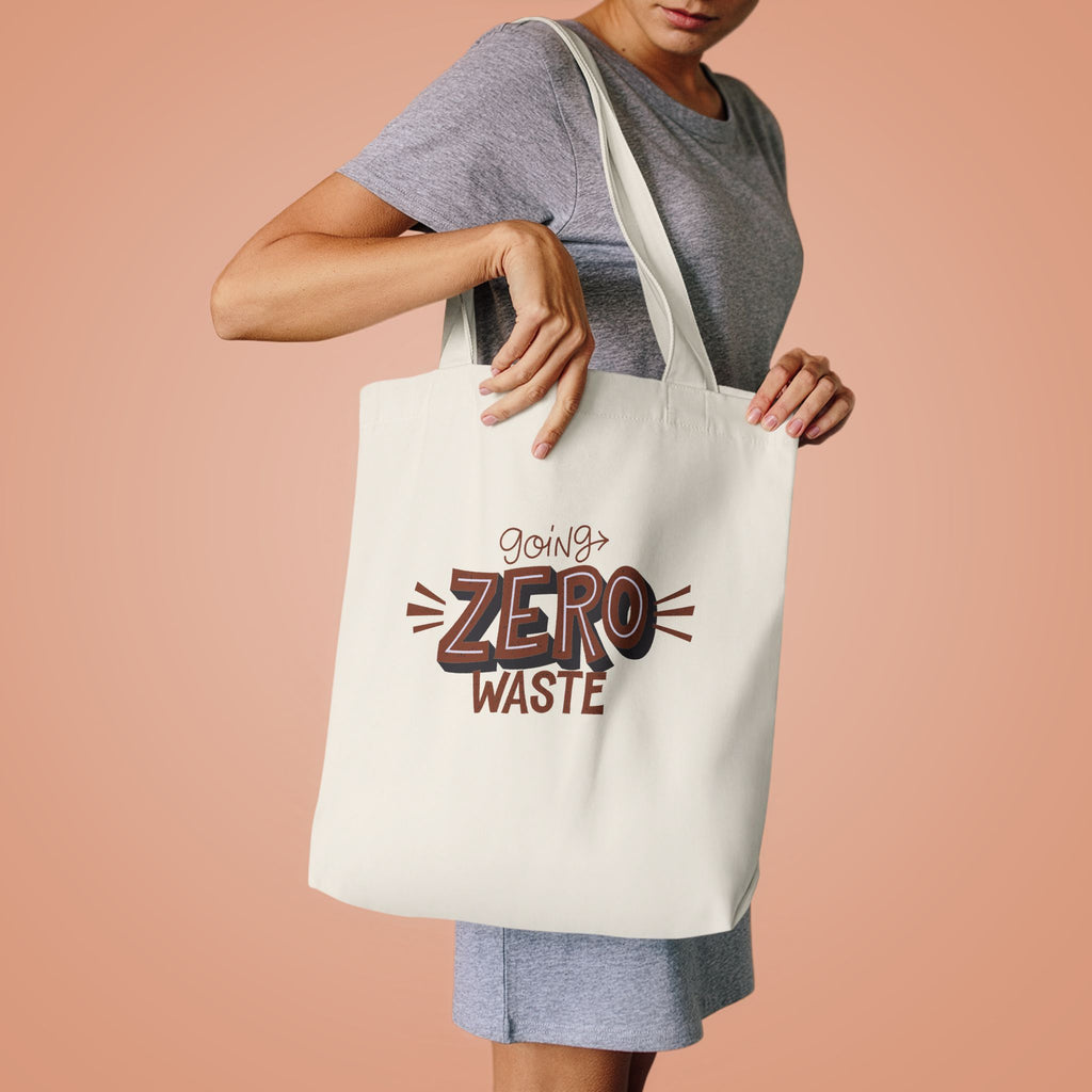 Woman in a grey tee lifting the Going Zero Waste tote by its handles against peach