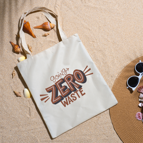 Going Zero Waste tote on beach sand with seashells, a straw hat and white sunglasses nearby